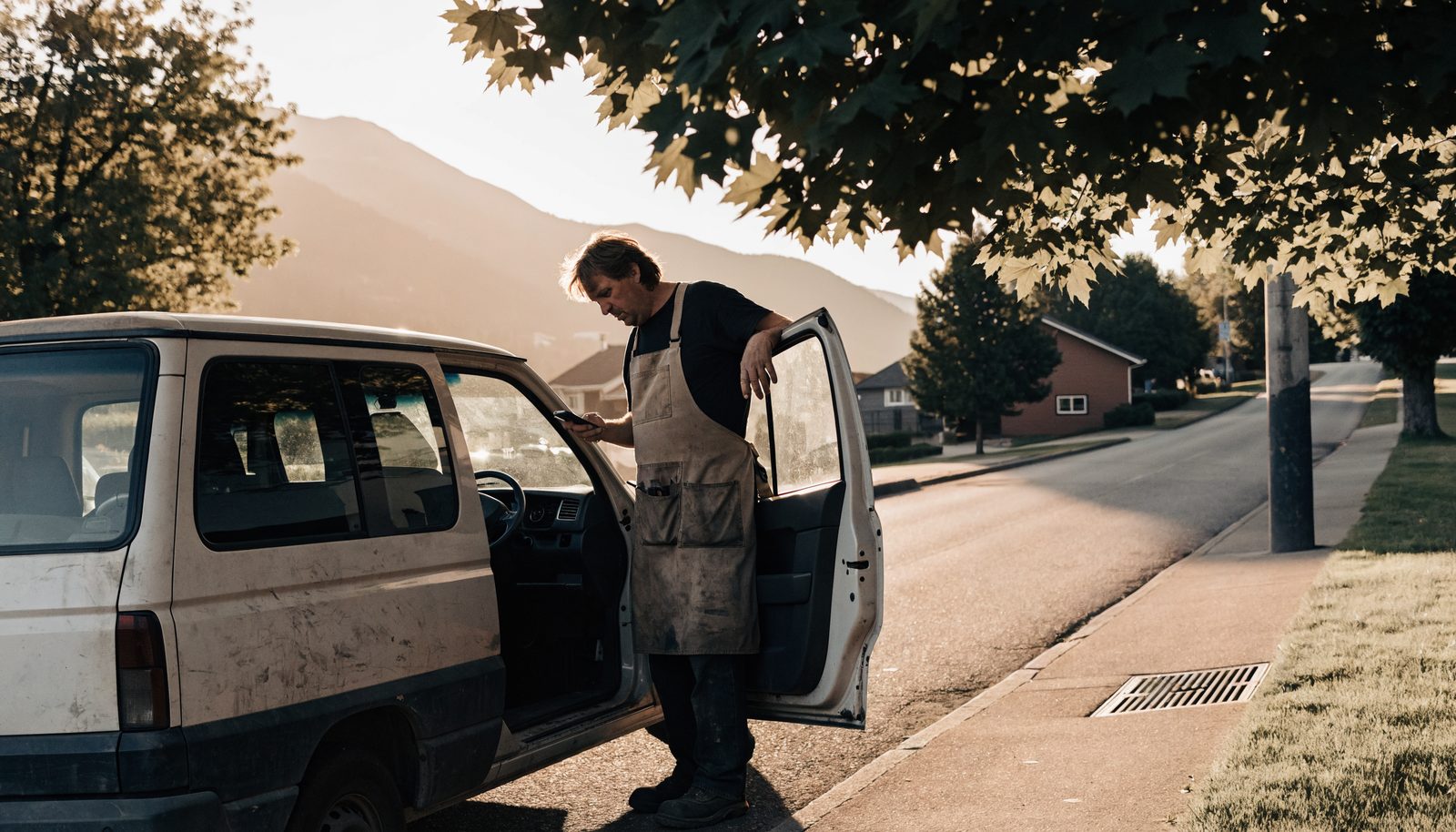 A tradesperson at an open work van on a quiet street, looking at a phone, mountains in the distance